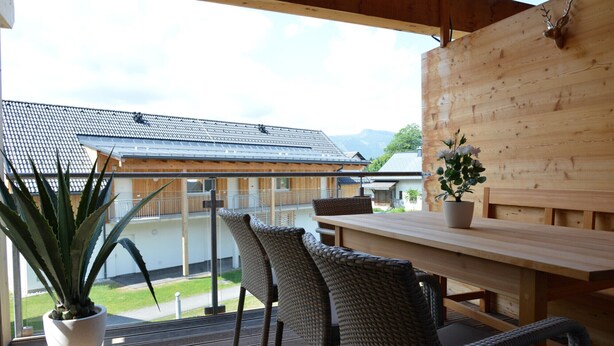 Una terraza de madera con sillas de mimbre y una mesa, con vistas a una montaña y una casa con tejado de tejas.