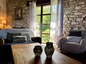 A room with a stone wall, a bed with a blue blanket, a wooden table with two vases, and a window with curtains. - 'Mas de la Laiterie' - Exceptional Provencal Charm between Luberon and Ventoux (Pernes-Les-Fontaines)