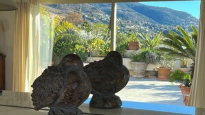 A view through a window with two bronze sculptures on a marble surface, overlooking a courtyard with potted plants and a mountainous landscape. - Magnifique appartement dominant de 100m2, énorme terrasse vue mer et montagne (Vence)