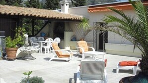 An outdoor patio with white lounge chairs and a table, surrounded by potted plants and a mountain backdrop. - Magnifique appartement dominant de 100m2, énorme terrasse vue mer et montagne (Vence)