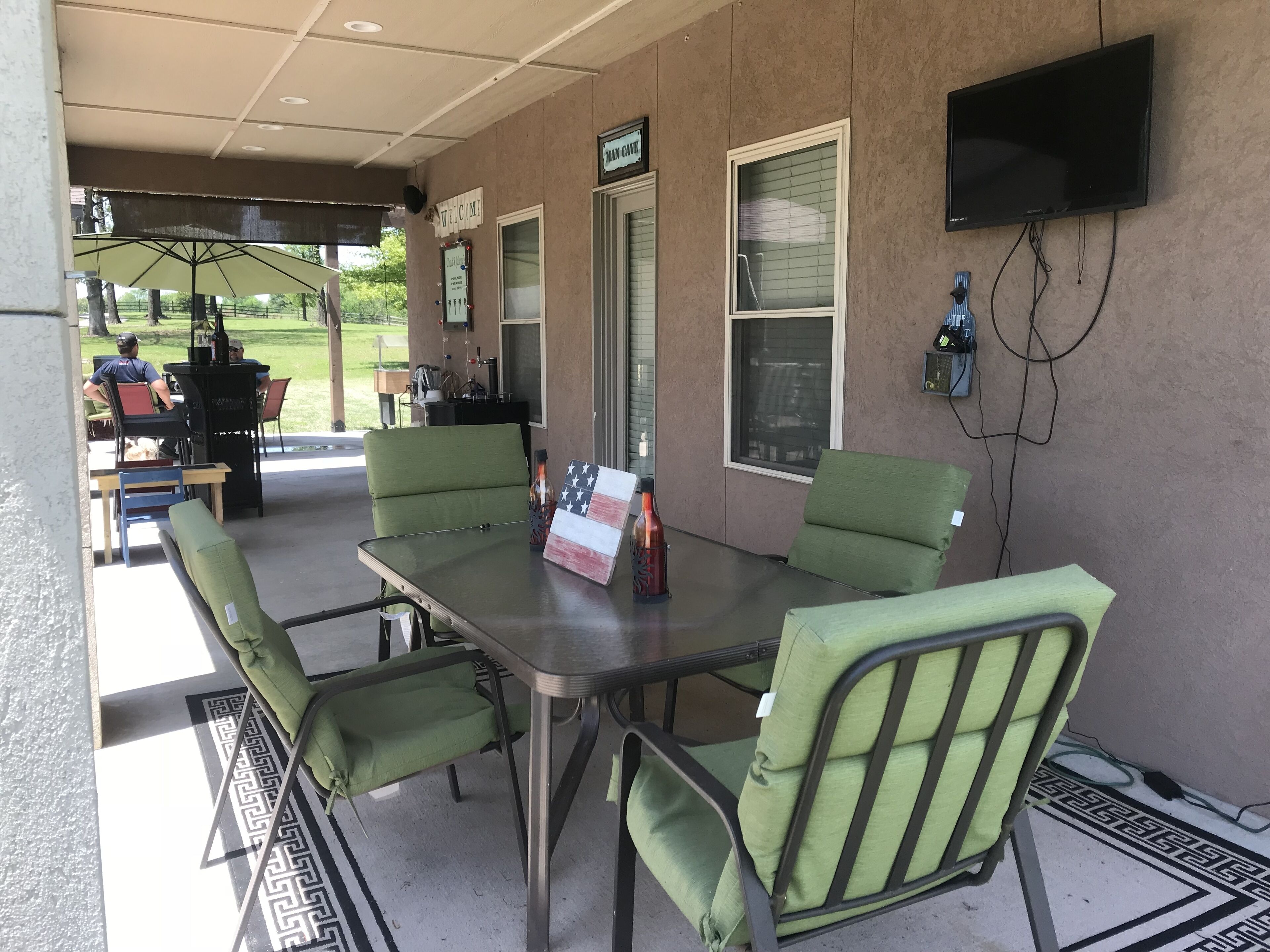 Un área de patio techada con mesa y sillas, un letrero de bienvenida y un televisor montado en la pared.