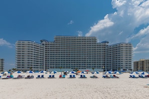 A large hotel with multiple floors and balconies, a sandy beach in the foreground with several beach chairs and umbrellas, and a clear blue sky.