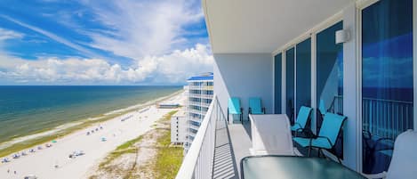 A balcony with a view of a beach and ocean, featuring a glass table and chairs.