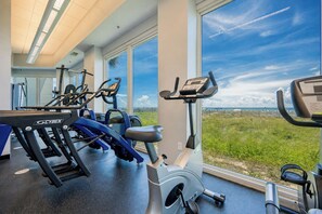 Uma academia com diversos equipamentos de exercício, janelas amplas e vista para a praia.