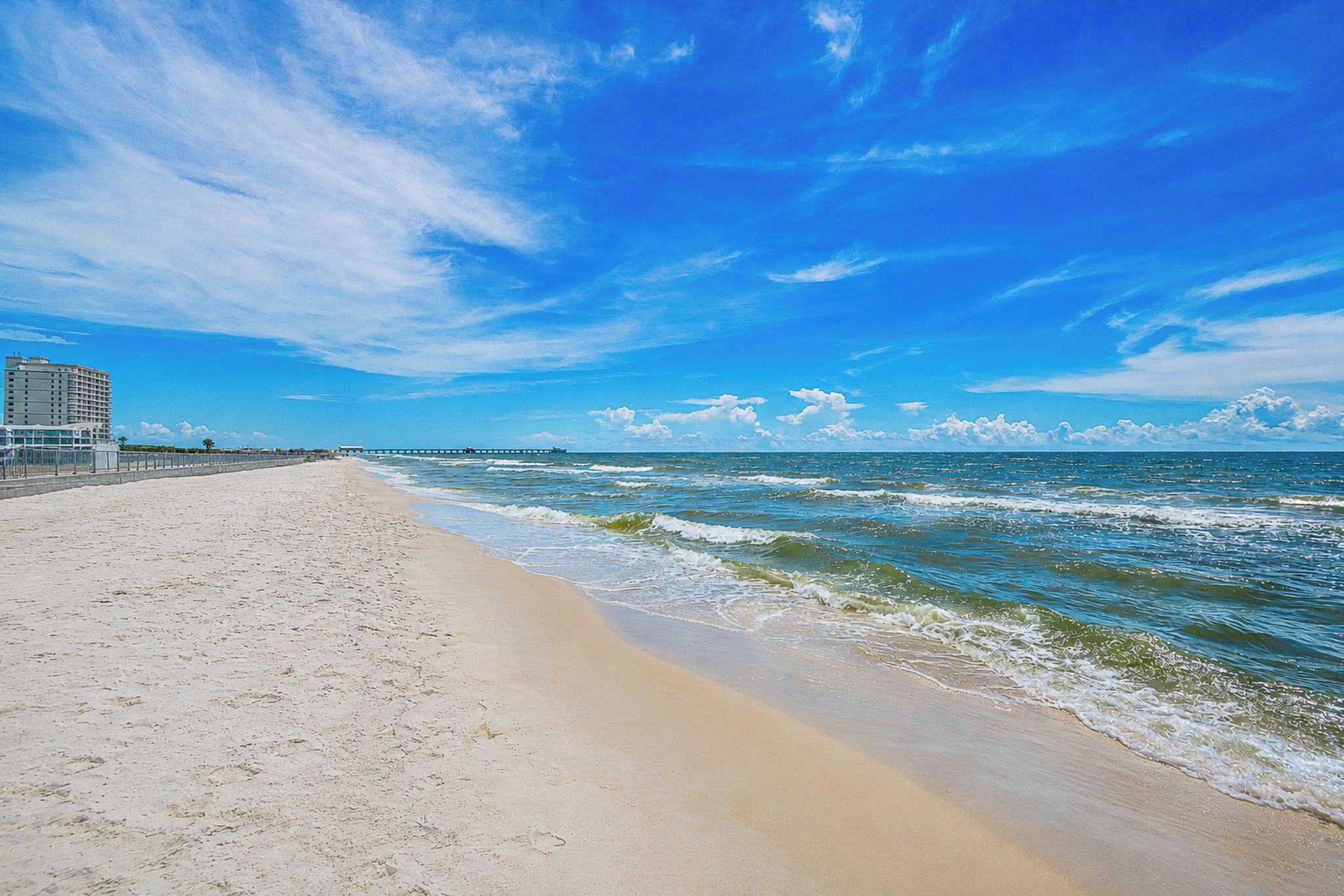 A beach with a clear sky, waves, and a pier in the distance.