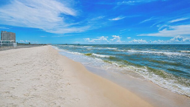 Een strand met een heldere lucht, golven en een pier in de verte.