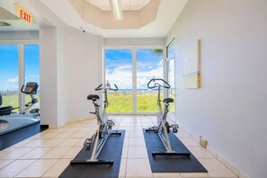 Un gimnasio con dos máquinas elípticas, una cinta de correr y un amplio ventanal con vista a la playa.