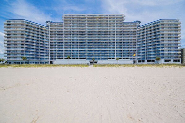 A large hotel with multiple floors and balconies, situated on a sandy beach.