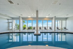 A large indoor swimming pool with clear blue water, surrounded by windows offering views of palm trees and a clear sky.