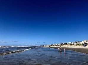 A beach with people walking along the shore, houses in the background, and clear blue skies.