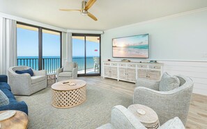 Ein geräumiges Zimmer mit großem Fenster zum Meer, einem Deckenventilator, einem Flachbildfernseher und einem Holzkasten.