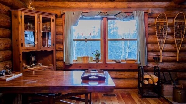 A rustic wooden cabin interior with a large dining table, a cabinet with glass doors, a window with curtains, and skis leaning against the wall.