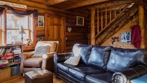 A cozy log cabin interior with a leather sofa, a cushioned armchair, and a wooden staircase.