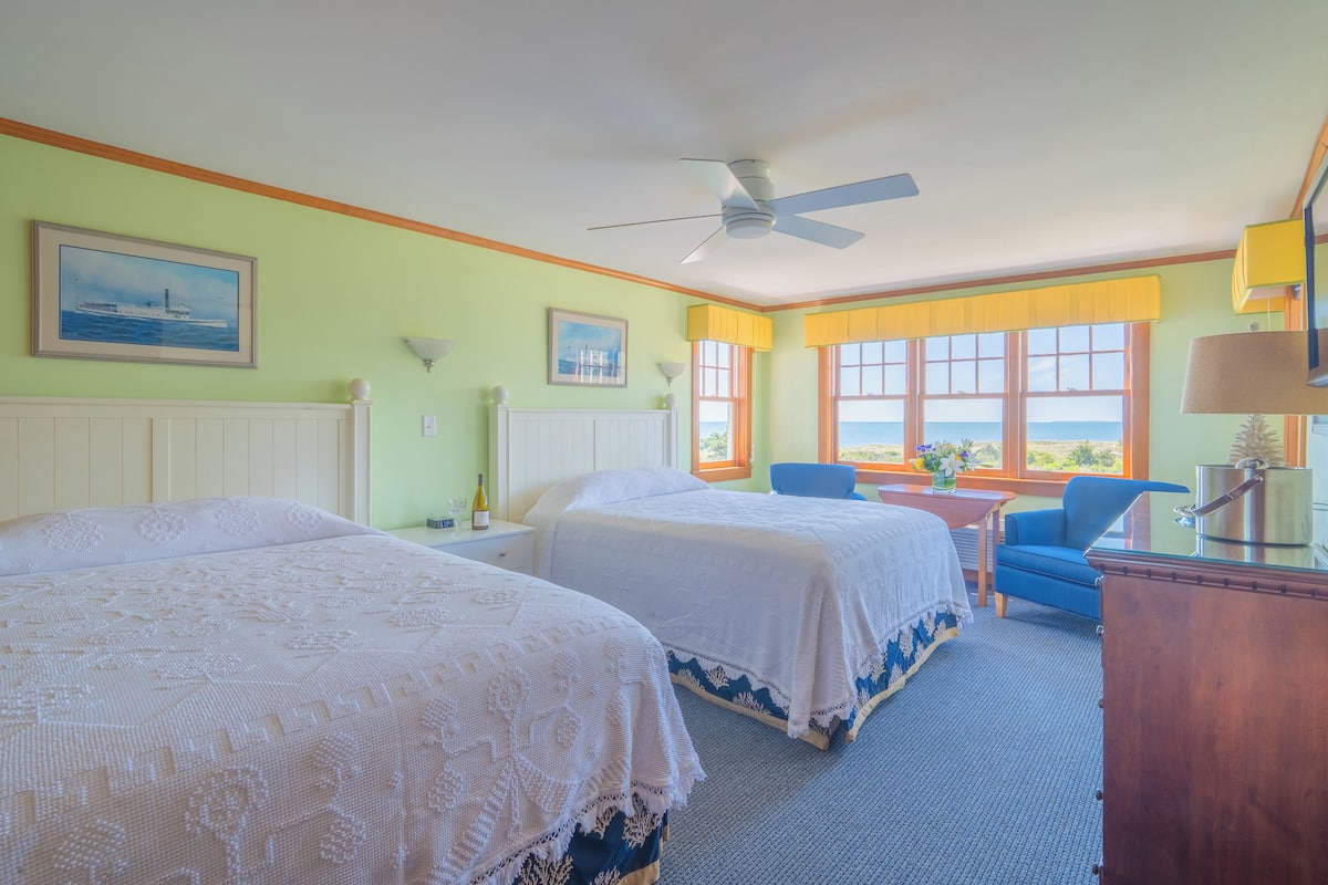 Une chambre d’hôtel avec deux lits, un bureau, une chaise, un ventilateur de plafond et de grandes fenêtres donnant sur un beau paysage.