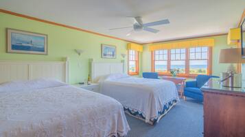 A hotel room with two beds, a desk, a chair, a ceiling fan, and large windows with a view.