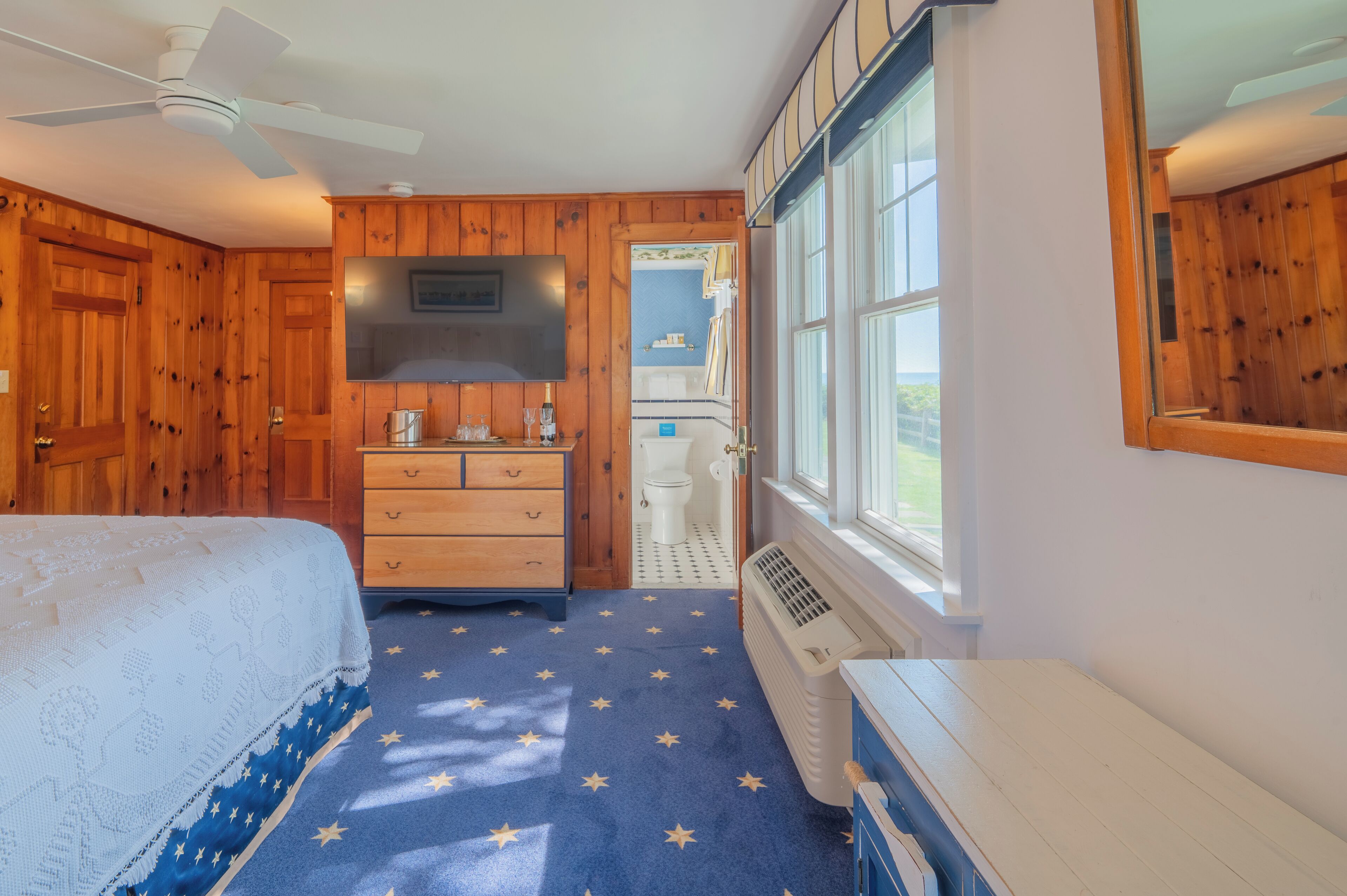 A bedroom with wooden paneling, a bed with a star-patterned rug, a dresser, a ceiling fan, and a window with a view.