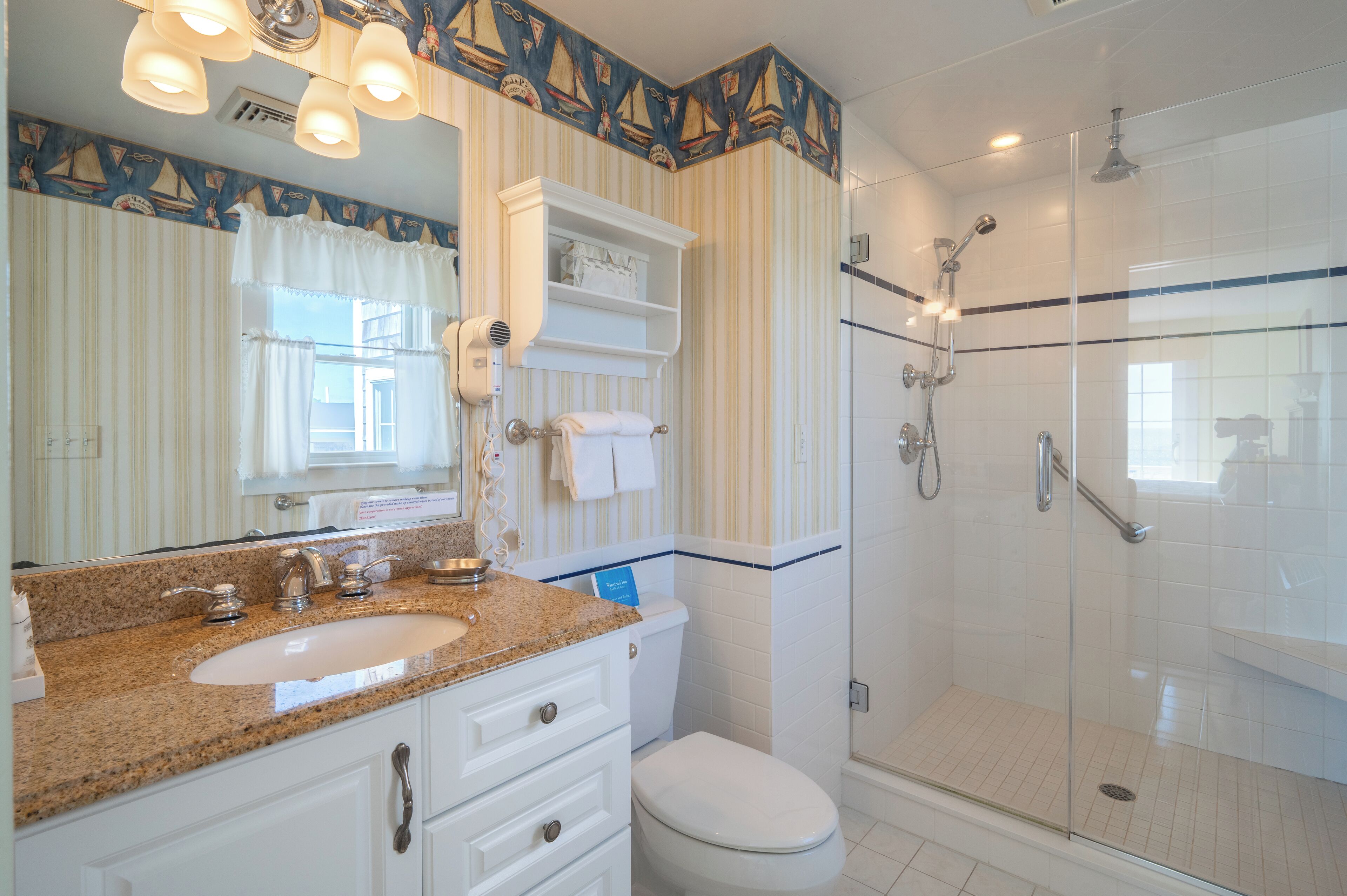 A bathroom with a granite countertop sink, a white toilet, a glass-enclosed shower, and striped wallpaper.