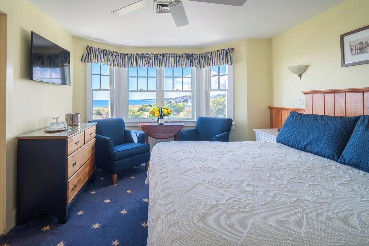Une chambre à coucher comprenant un lit, deux chaises, une commode, une table de chevet, une lampe, une fenêtre avec vue et un ventilateur de plafond.