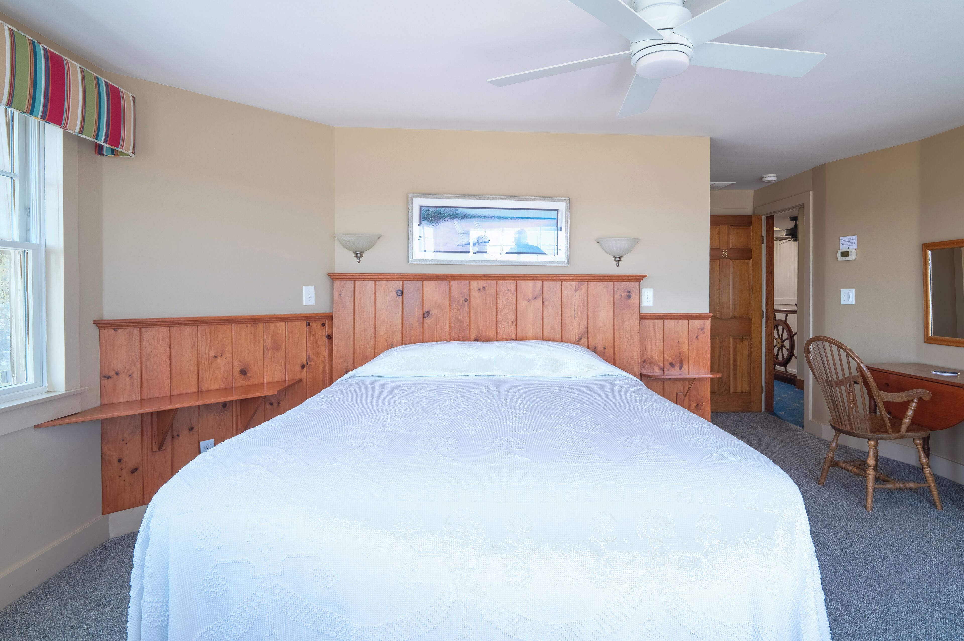 A bedroom with a large bed, wooden headboard, a ceiling fan, a window with curtains, and a chair.