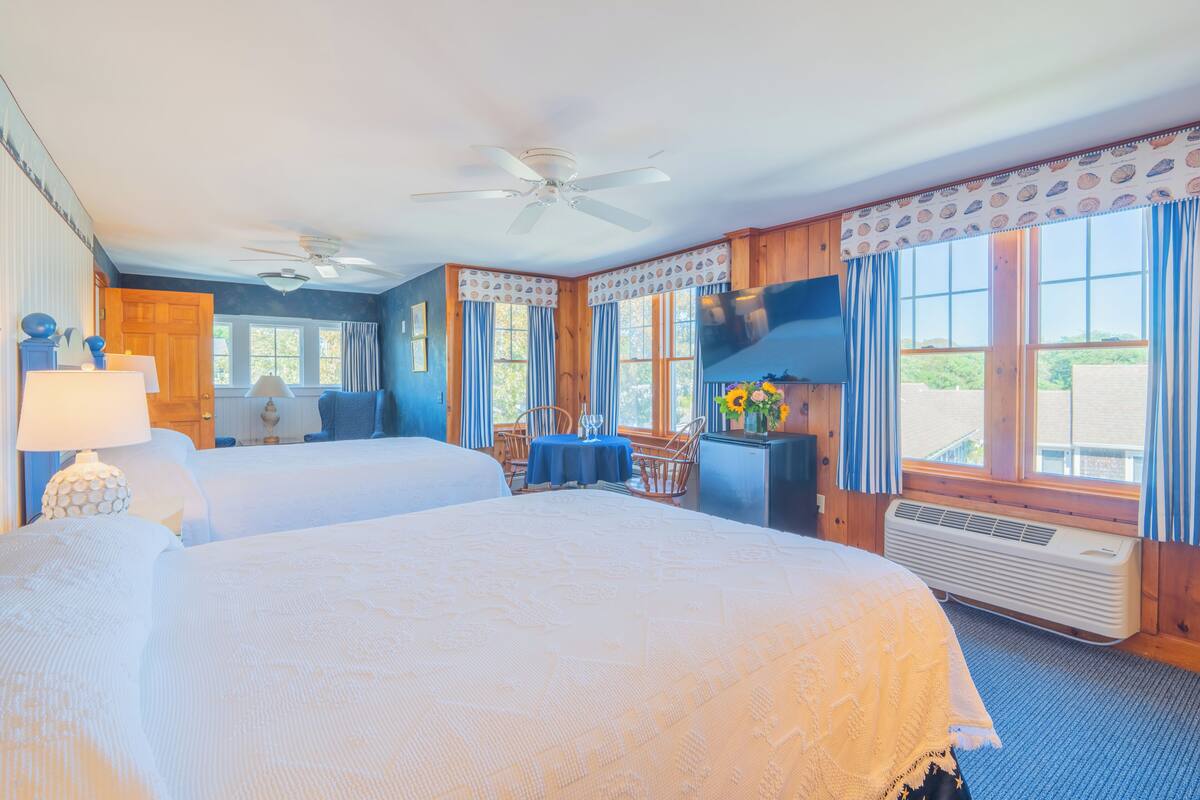 Une chambre d’hôtel avec deux lits, un ventilateur de plafond, une grande fenêtre avec des rideaux, une table avec des chaises et un petit bureau avec une lampe.