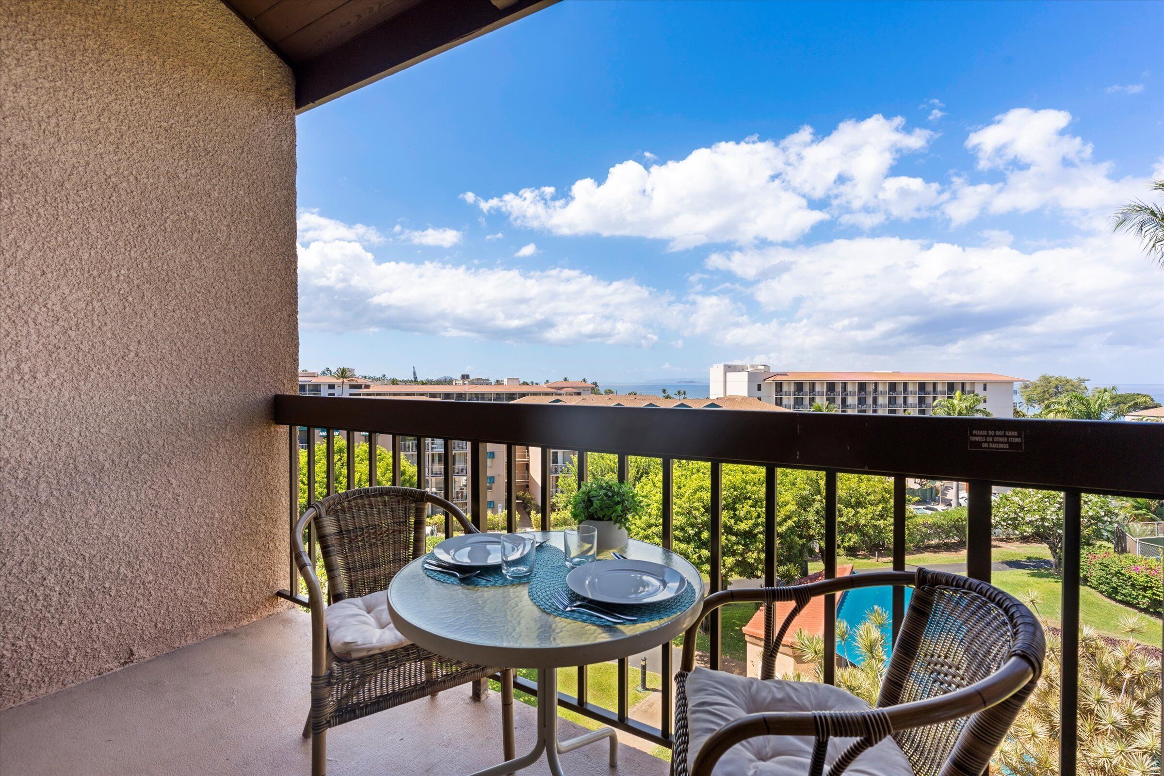 Un balcon avec des fauteuils en osier, une table en verre mise avec des assiettes et des verres, et une vue sur une piscine et des bâtiments en arrière-plan.