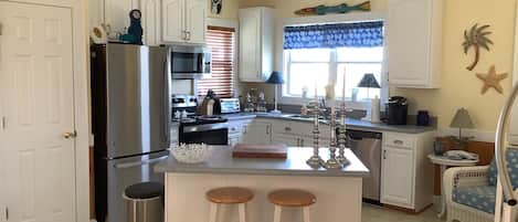 A kitchen with white cabinets, a stainless steel refrigerator, a microwave, a sink, and a window with blinds. There are two stools in front of the counter.