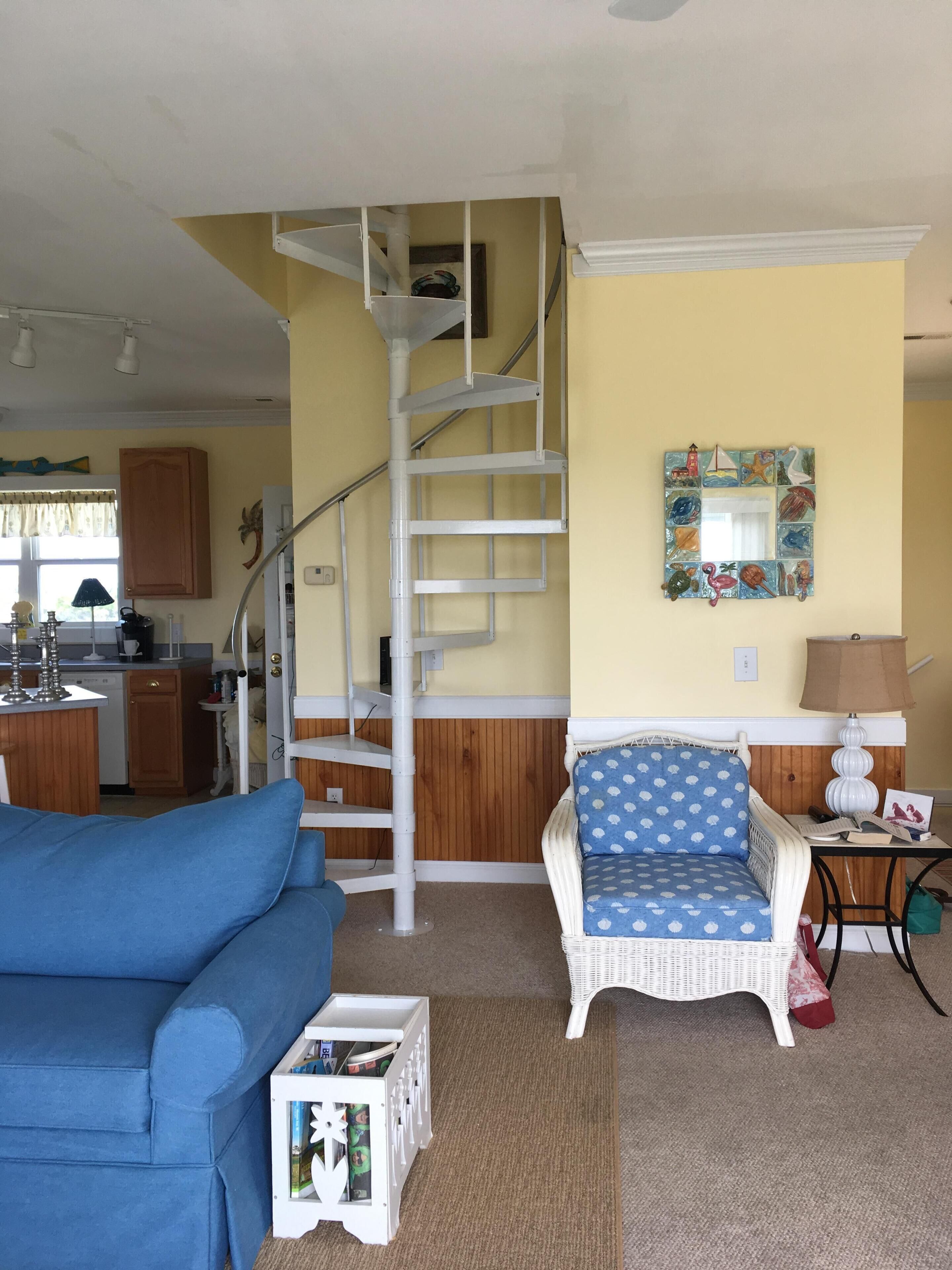 Una sala de estar con un sofá azul, una silla de mimbre, una escalera en espiral blanca y una cocina al fondo.