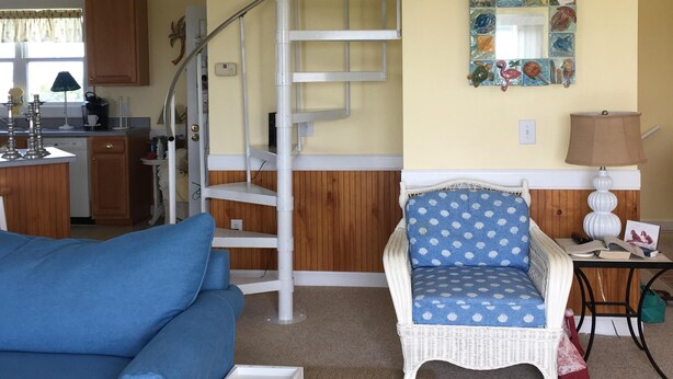 Una sala de estar con un sofá azul, una silla de mimbre, una escalera en espiral blanca y una cocina al fondo.