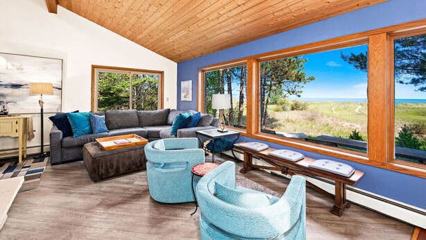 Un salón moderno con techo de madera, ventanas amplias, un sofá gris, sillas azules y vistas al exterior.