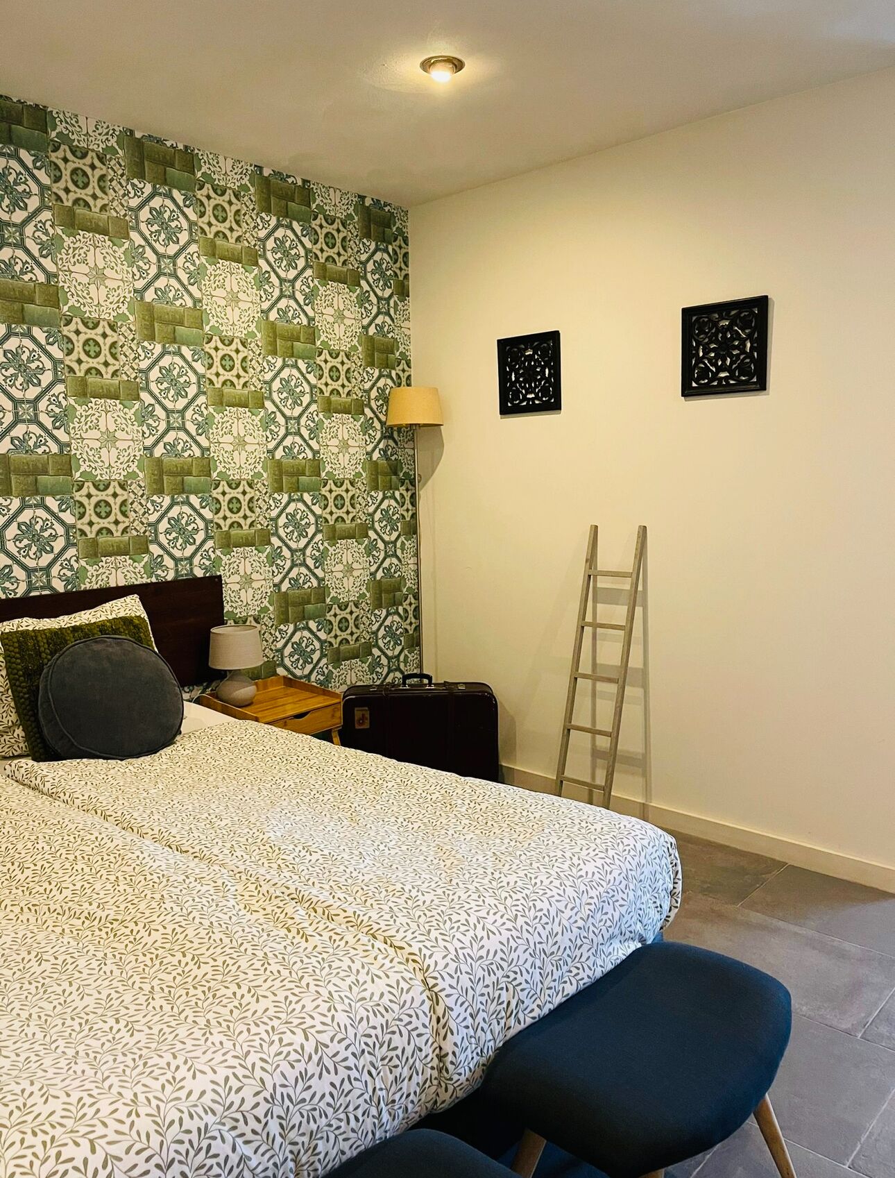 A neatly arranged bedroom with a patterned bedspread, a bedside lamp, a suitcase, and a ladder against the wall.