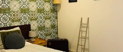 A neatly arranged bedroom with a patterned bedspread, a bedside lamp, a suitcase, and a ladder against the wall.