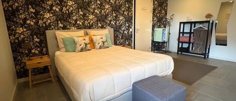 A bedroom with a bed, a bedside table, a grey ottoman, a wardrobe, and a bathroom door.