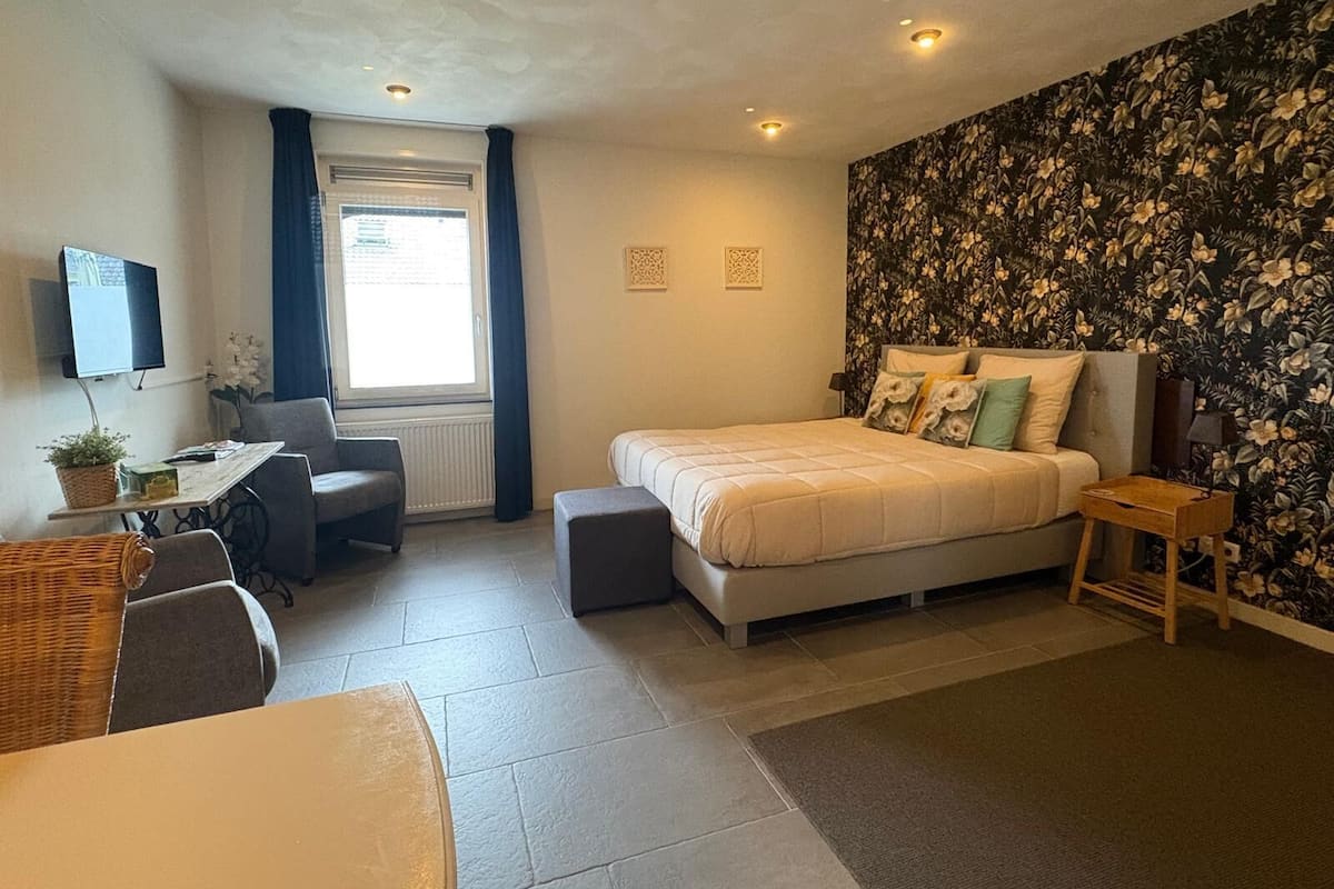 A bedroom with a bed, a desk, a chair, a TV, and a window.