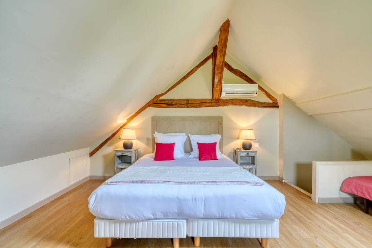 A bedroom with a sloped ceiling, a large bed with white bedding and red pillows, two bedside tables with lamps, and a radiator on the wall.