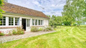 A white building with a tiled roof, a brick foundation, and a small garden with a table and chairs.