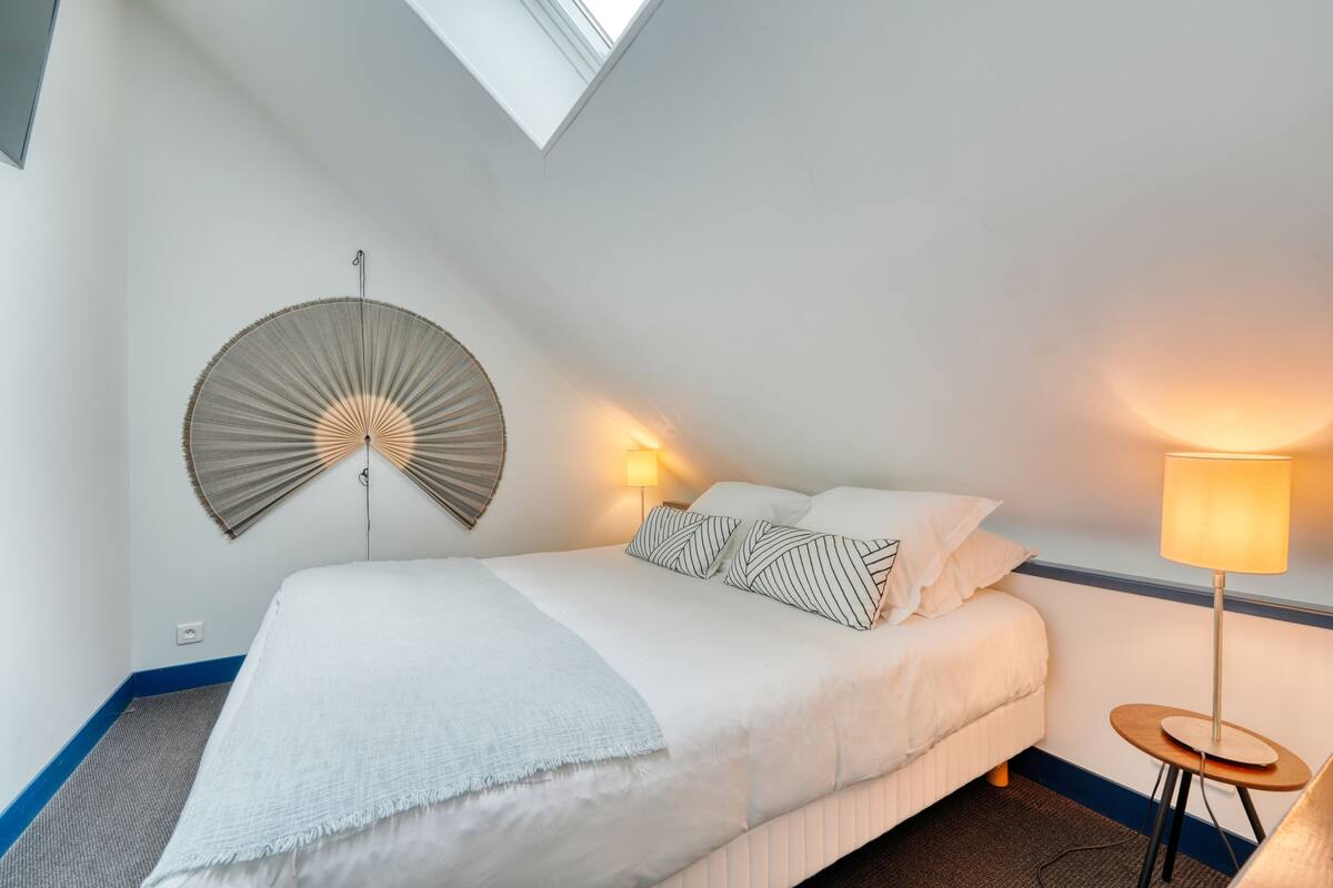 A hotel room with a bed, a wall-mounted fan, a bedside lamp, and a small table.