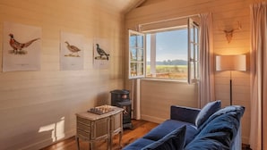 Una habitación acogedora con techo de madera, un ventanal con cortinas, un sofá azul, una mesita con un tablero de ajedrez e ilustraciones de pájaros en la pared.