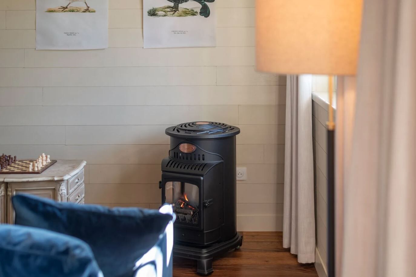 A cozy room with a wood-burning stove, a chessboard on a dresser, and a lamp.
