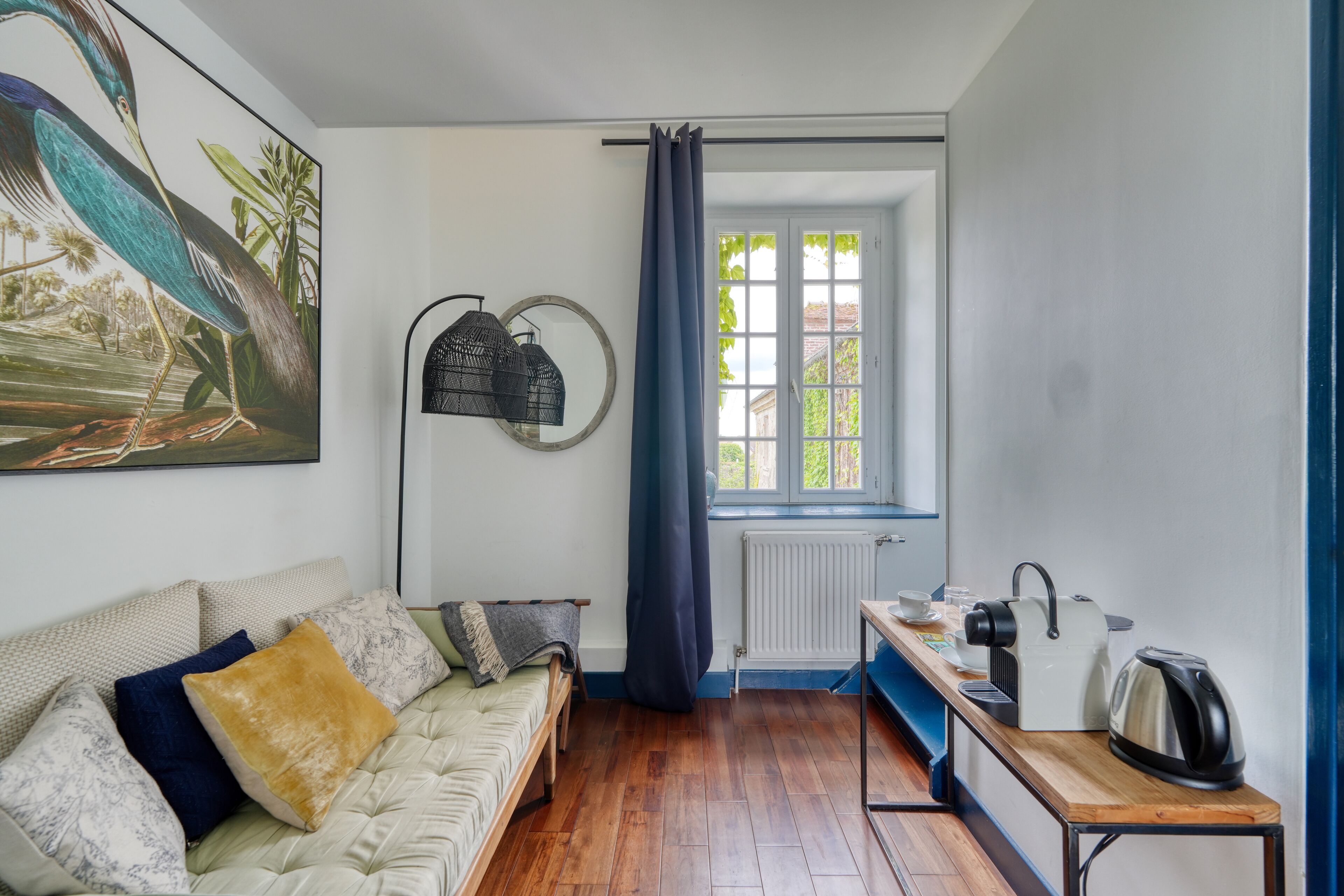 A cozy room with a sofa, a small table with a kettle, a window with curtains, and a wall-mounted lamp.