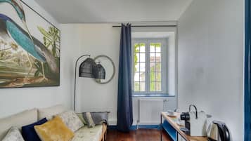 A cozy room with a sofa, a small table with a kettle, a window with curtains, and a wall-mounted lamp.