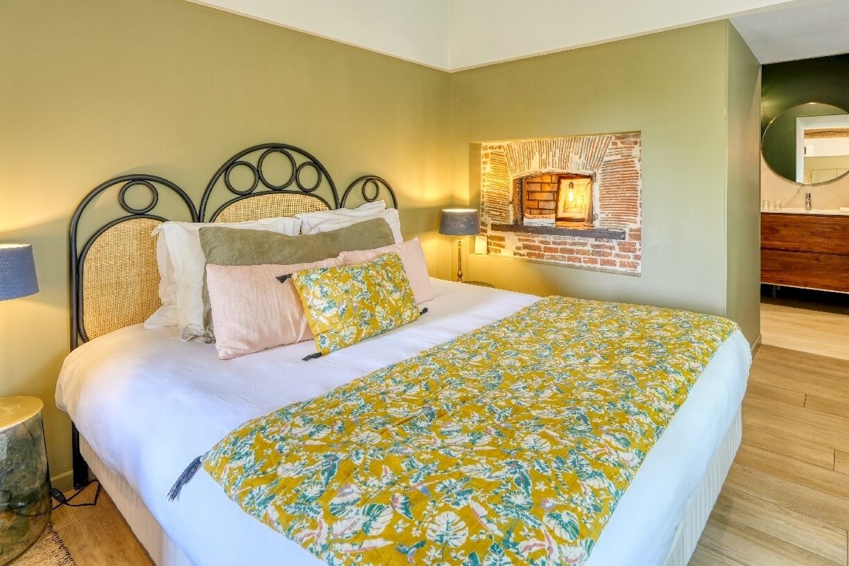 A bedroom with a bed, a lamp, a wall-mounted fireplace, and a wooden dresser.