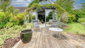 Una terraza de madera con dos sillas y una mesa, rodeada de exuberante vegetación y una casa con techo verde.