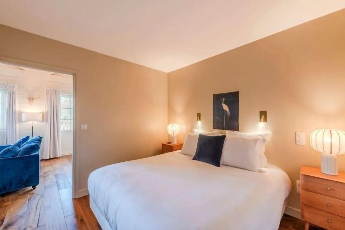 A bedroom with a large bed, bedside lamps, a nightstand, and a door leading to another room.