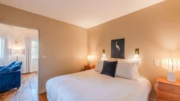 Une chambre à coucher avec un grand lit, deux tables de chevet avec des lampes, un fauteuil bleu et une porte donnant sur une autre pièce.