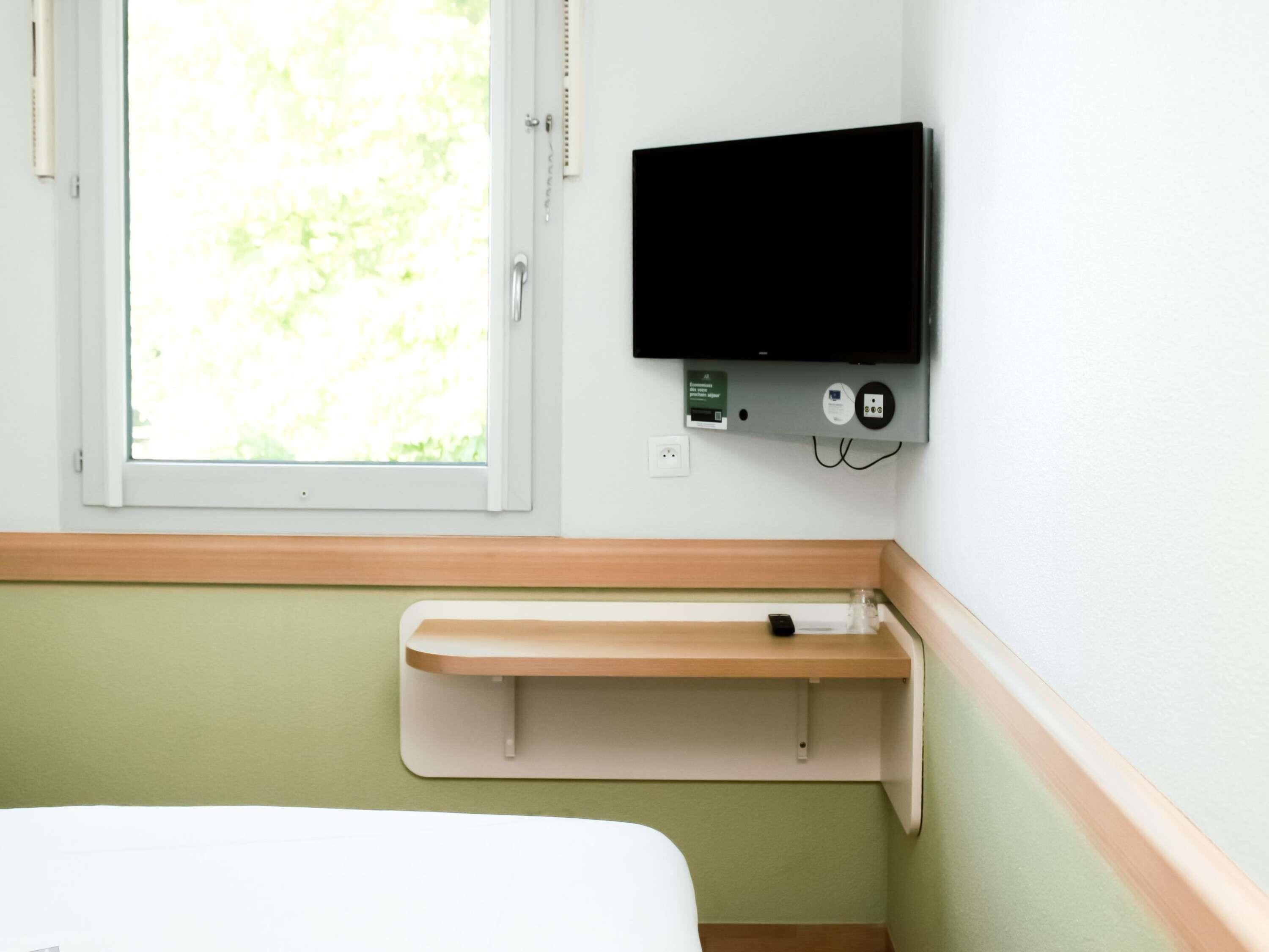 A hospital room with a bed, a window with a view of greenery, a mounted TV, and a small shelf.