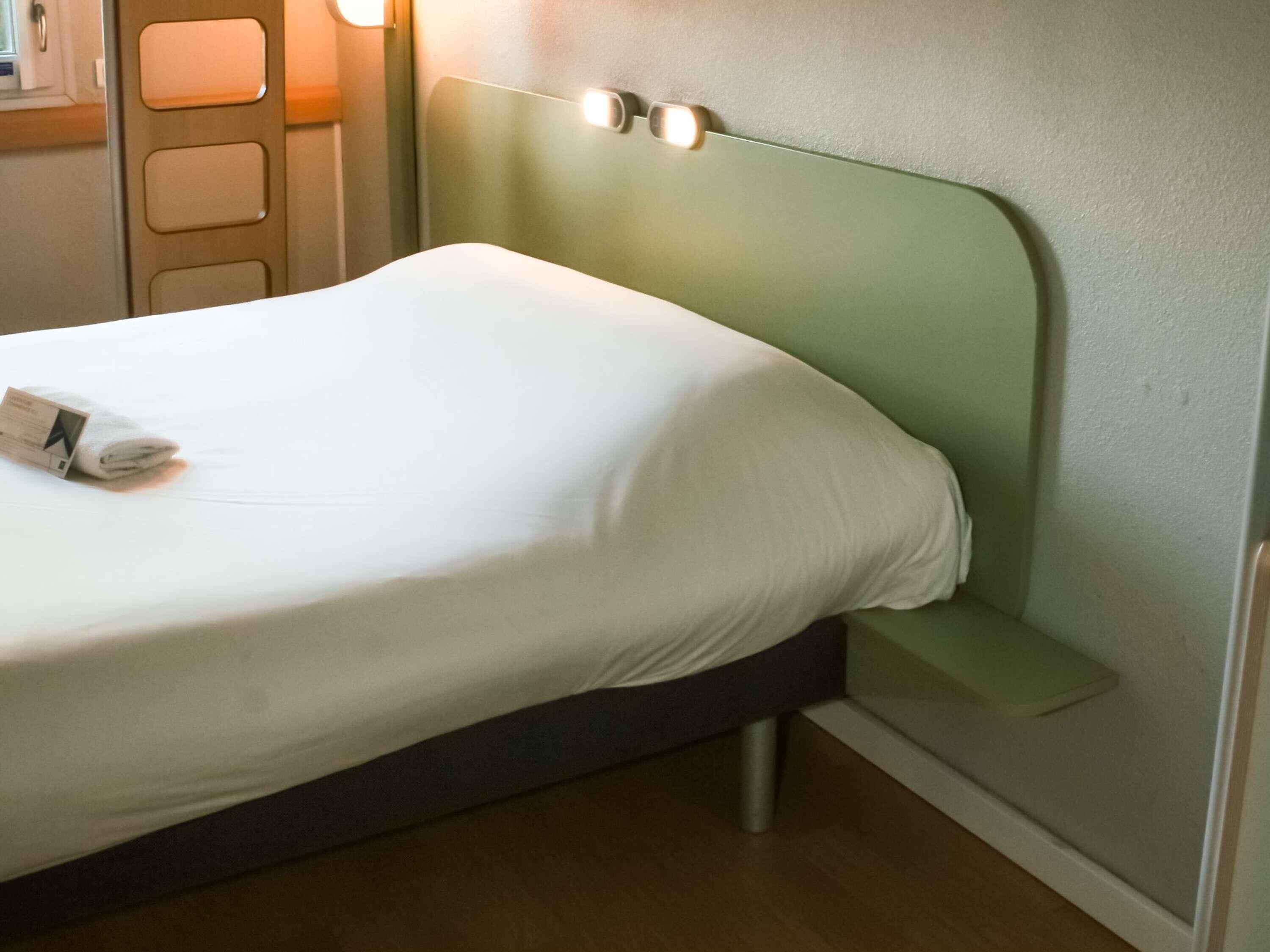 Un lit bien fait, avec un drap blanc et une serviette pliée, une tête de lit verte dotée de deux interrupteurs et un plancher en bois.