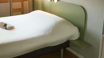 Un lit bien fait, avec un drap blanc et une serviette pliée, une tête de lit verte dotée de deux interrupteurs et un plancher en bois.