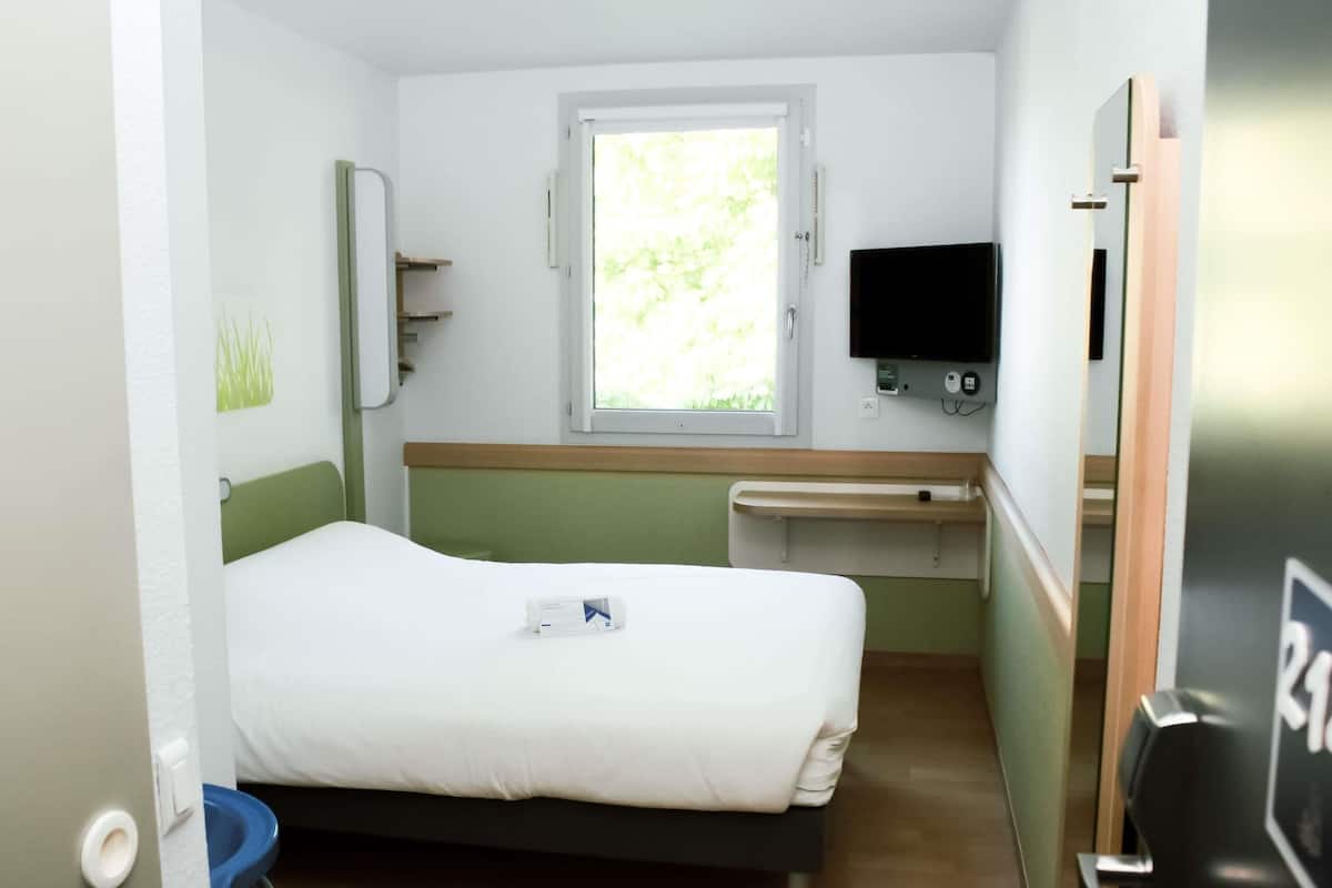 Ein Einzelbett mit weißer Matratze, ein kleiner Nachttisch, ein Fenster mit Blick ins Grüne, ein flachbildschirmfernseher an der Wand und eine Tür mit Griff.