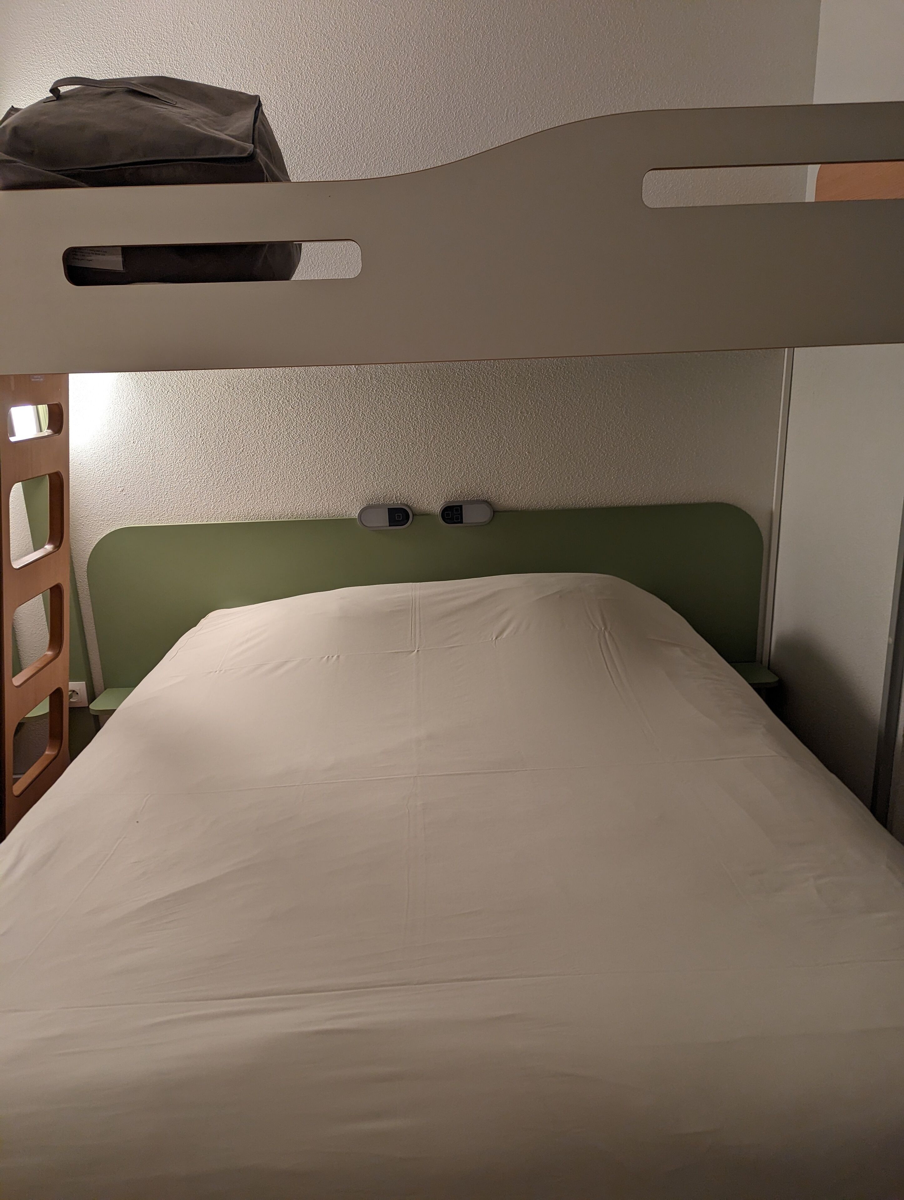 Una cama litera con cabecera verde y colchón blanco.  A la izquierda, hay una mesita pequeña y una escalera.