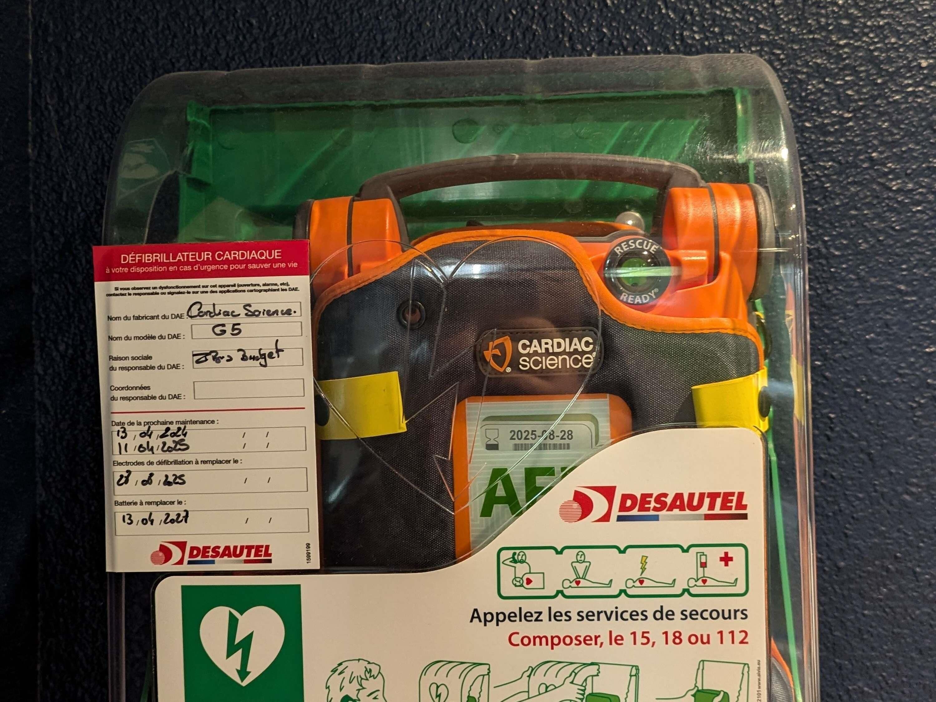 A defibrillator in a plastic case with a maintenance label and emergency instructions.